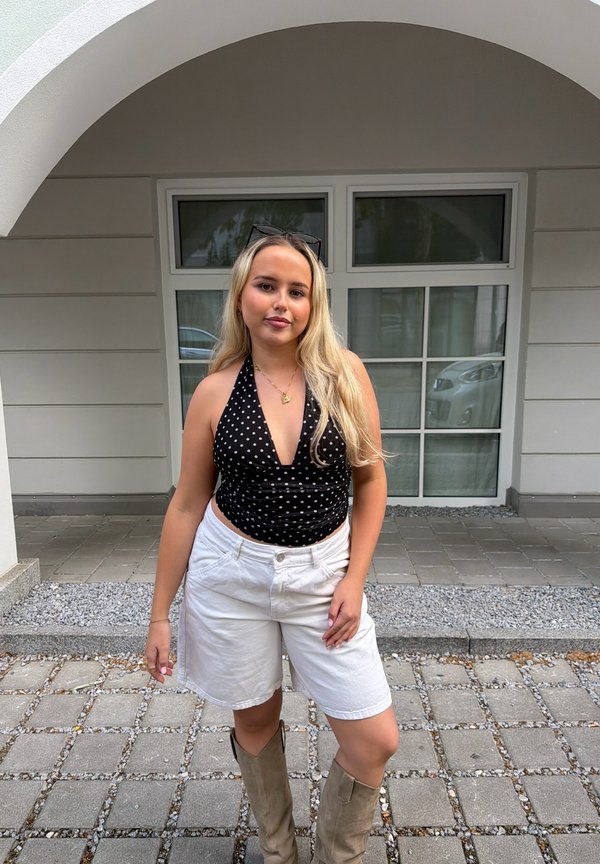 Jeune femme aux longs cheveux blonds portant un haut noir à pois, un short blanc et des bottes beiges, debout devant un bâtiment moderne.
