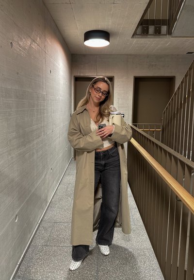 Abrigo largo beige sobre top blanco y jeans negros, complementado con zapatos blancos que presentan detalles perforados. El fondo incluye paredes grises y una barandilla.