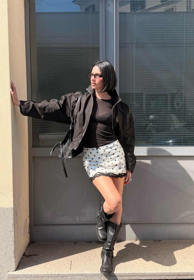 Naine musta bob-soenguga, kellel on seljas must jakk, läbipaistev pluus, valge täpiline seelik ja mustad saapad, toetumas seinale päikesevalguses.