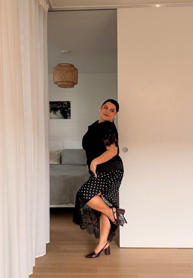 Femme en haut noir et jupe à pois levant une jambe, debout dans l'encadrement d'une chambre avec des rideaux blancs et un sol en bois.