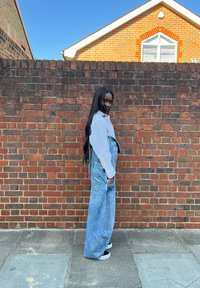 Modre jeans salopete s širokimi hlačnicami in stranskimi žepi, oblečene čez belo srajco z dolgimi rokavi, stojijo ob zidani steni.