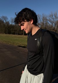 Camisa negra de manga larga con un pequeño logo blanco, pantalones cortos gris claro y una mochila negra, de pie sobre un suelo pavimentado con césped y árboles.