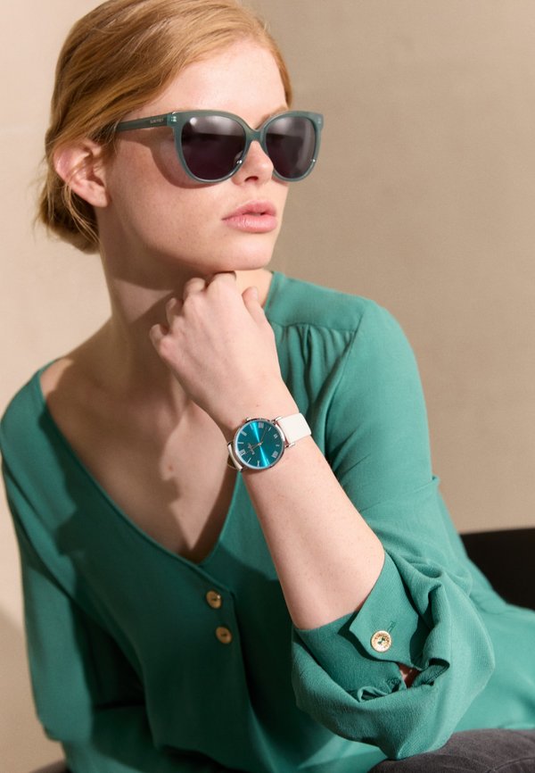 Grüne Bluse mit Knopf-Akzenten, türkisfarbene Uhr mit weißem Armband, übergroße türkisfarbene Sonnenbrille, glatte Textur, im Sitzen vor einem neutralen Hintergrund.
