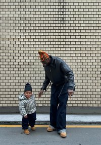 En vuxen i en svart läderjacka och en färgglad randig mössa står bredvid ett barn i en rutiga hoodie och svarta byxor, båda bärande bruna skor.