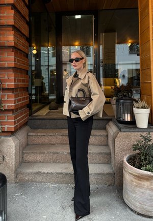 Mujer con chaqueta corta beige, pantalones negros y gafas de sol, sosteniendo un bolso negro, de pie en las escaleras frente a la entrada de un edificio.