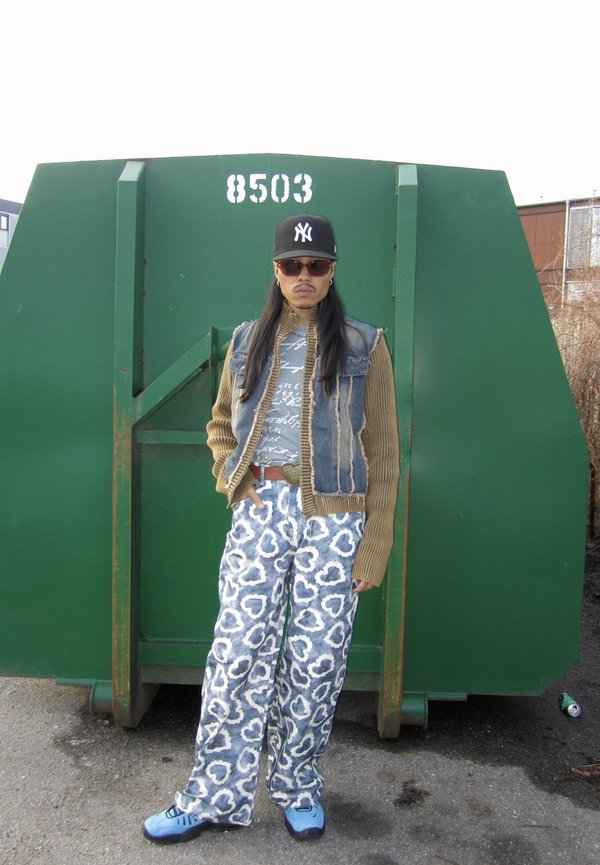 Gilet en denim sur une chemise grise à motifs, pantalon à motifs de cœurs bleu et blanc, chaussures bleu clair, se tenant devant une benne à ordures verte.