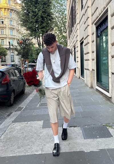 Una persona lleva una camiseta gris, pantalones cortos beige y zapatos negros. Sostiene un ramo de rosas rojas y tiene un suéter marrón colgado sobre los hombros.