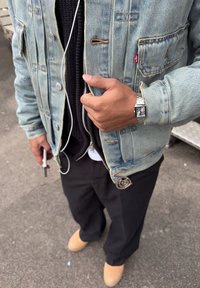 Personne portant une veste en denim clair, un pull noir, un pantalon noir, des chaussures marron clair, une montre argentée, tenant un téléphone avec des écouteurs blancs branchés.