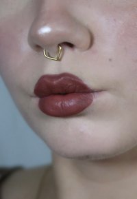 Anello al setto in oro con forma geometrica, rossetto opaco marrone scuro su labbra piene, texture della pelle liscia e leggero luccichio sul naso.