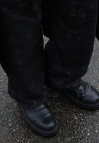 Chaussures en cuir noir portées avec un pantalon noir ample, debout sur un sol en gravier humide.