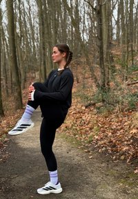 Chemise noire à manches longues, leggings noirs, chaussures de sport légères avec des rayures noires, et chaussettes à rayures violettes ; en plein air sur un sentier entouré d'arbres.