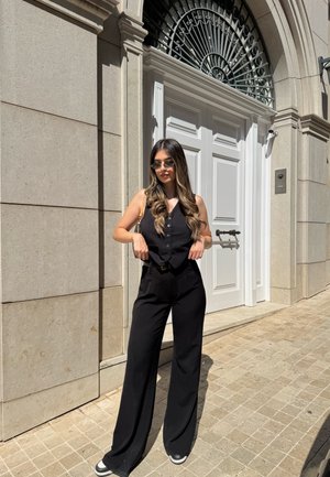 Mujer con cabello largo y castaño que lleva una camiseta sin mangas negra, pantalones de pierna ancha y gafas de sol, de pie sobre un pavimento de piedra iluminado por el sol junto a unas puertas dobles blancas.