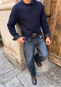 Maglione navy a coste sopra una camicia azzurro chiaro, abbinato a jeans di denim scuro larghi e stivali in pelle marrone, in piedi contro un muro di pietra.