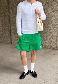 Weißes Langarmhemd, grüne Shorts, schwarze Slipper mit weißen Socken, hält eine kleine weiße Handtasche. Die Shorts haben ein Logo-Detail.