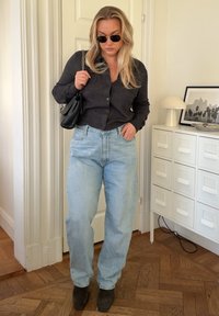 Svart cardigan med knapper, lyseblå høyhøylivet jeans, grå ankelstøvler og svart håndveske; solbriller fullfører antrekket.