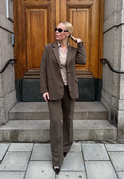 Blazer marrón ajustado sobre una blusa beige, combinado con pantalones de campana marrones. Gafas de sol delgadas y tacones puntiagudos completan el look.