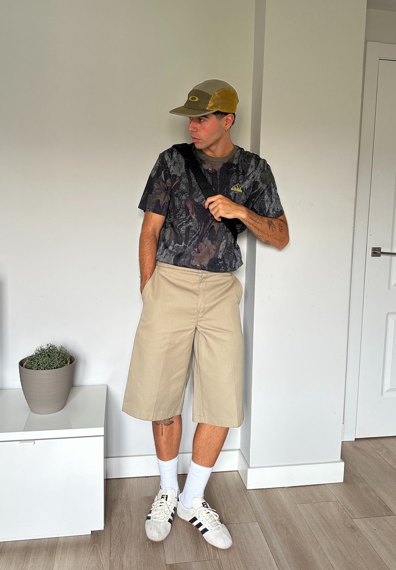 Chemise à manches courtes avec un motif camouflage, shorts beiges jusqu'aux genoux, chaussettes blanches et baskets blanches avec des rayures noires. Fond neutre avec une plante.