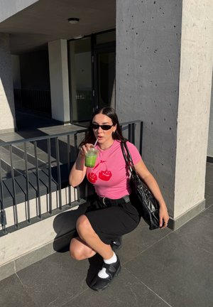 Mujer con gafas de sol, camiseta rosa con cerezas, falda negra y zapatillas deportivas, en cuclillas mientras bebe zumo verde afuera cerca de una pared de hormigón y una barandilla.