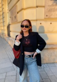 Μαύρο σακάκι πάνω από ένα κοντό μαύρο τοπ με κόκκινες λεπτομέρειες, ανοιχτό μπλε τζιν ψηλής μέσης, μαύρη ζώνη, κόκκινη τσάντα και γυαλιά ηλίου.