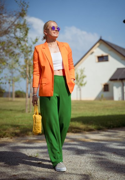 Americana naranja, top corto blanco, pantalones verdes holgados, zapatillas de cuadros en blanco y negro, bolsa tejida amarilla, entorno al aire libre con árboles.