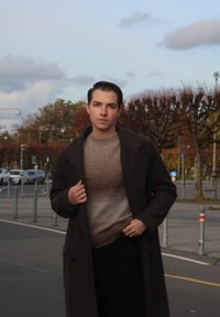 Lang brun overcoat båret over en tekstureret, gradient brun sweater. Personen står på en gade med træer og parkerede biler i baggrunden.