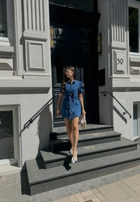  džinsinis suknelė, su pūstomis rankovėmis, diržu, ir apykakle. Dėvimi pilki batai. Laiptai yra iš tamsios akmens, apsupti baltos architektūros.