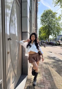 Camisa blanca de botones recortada, falda marrón con volantes y acentos de anillo metálico, botas negras hasta la rodilla, de pie junto a una puerta en un camino de piedra.