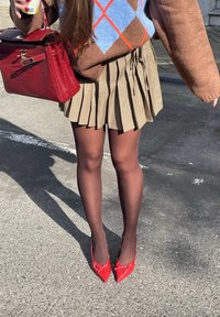 Bruine trui met een blauw en oranje ruitpatroon, geplooide rok in een ruitmotief, sheer zwarte panty, en rode puntige hakken met strikaccenten. Rode handtas.