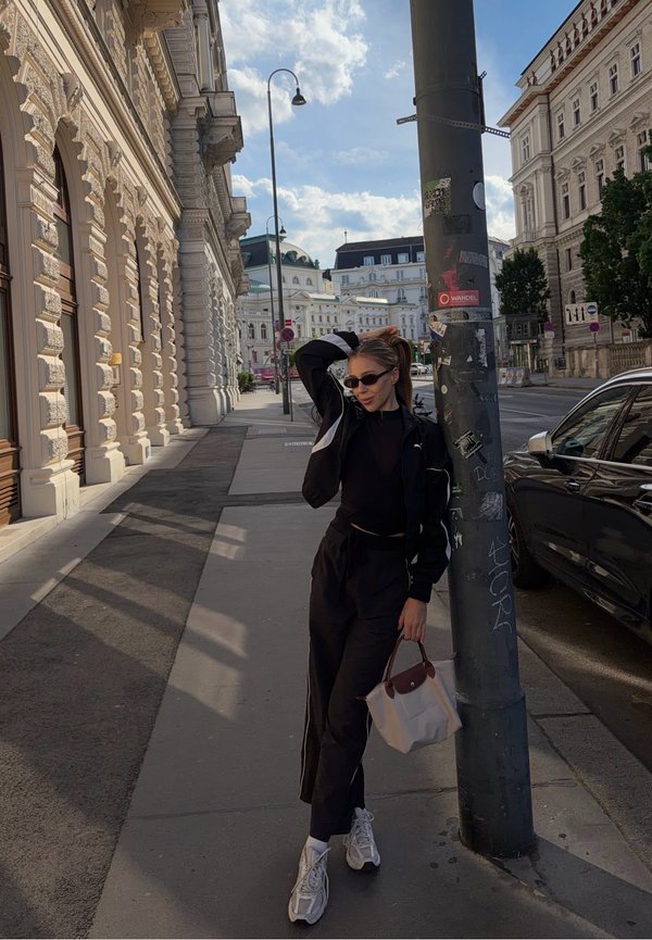 Jeune femme en tenue de sport noire et lunettes de soleil appuyée sur un lampadaire sur un trottoir en ville avec des bâtiments historiques et une voiture à proximité.