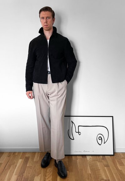 Chaqueta de ante negra con cremallera y cuello alzado, pantalones plisados beige y mocasines negros. Obra de arte abstracta enmarcada en el suelo detrás.