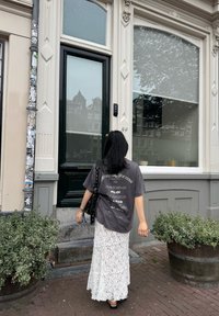 Grijze oversized graphic t-shirt met witte tekst, gecombineerd met een gelaagde witte kanten rok. Donkere handtas, zwarte sandalen en groene bladeren in de buurt.