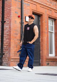 Črna brezrokavnika s roza logotipom, modri jeans in bele superge. Klobuk je oranžen z črnim logotipom. Gladka opečna stena v ozadju.