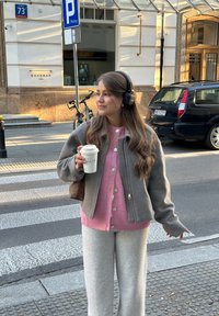 Jonge vrouw met een koptelefoon en een takeaway koffiekop staat nabij een zebrapad op een straat in de stad.