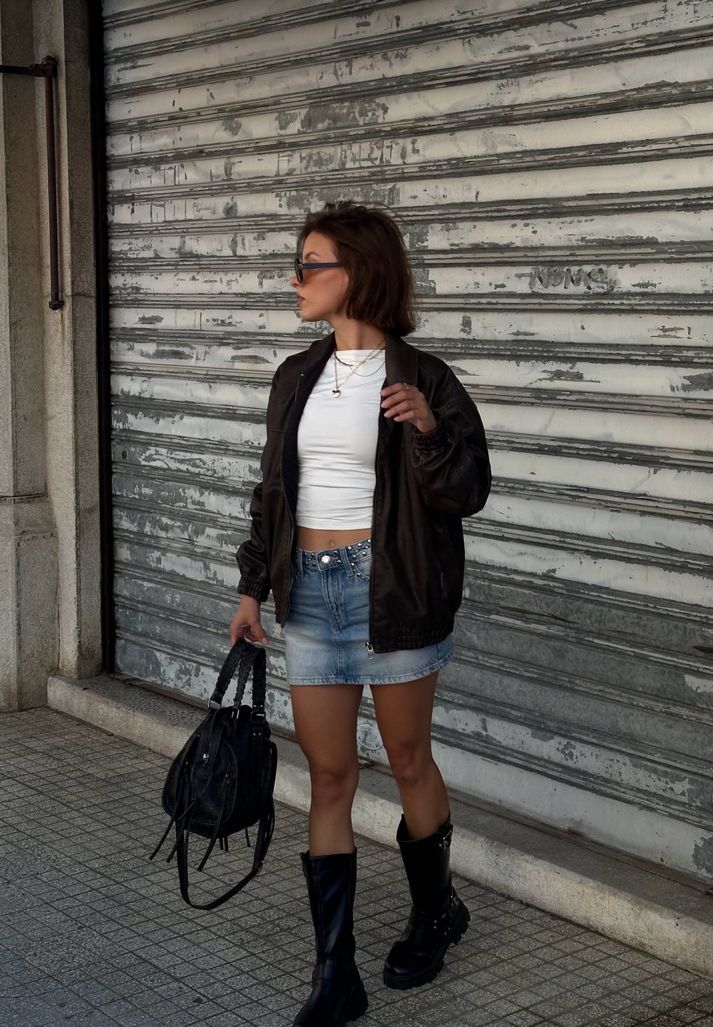 Frau in schwarzer Jacke, weißem Croptop, Denim-Minirock, schwarzen Stiefeln und Sonnenbrille, die eine schwarze Handtasche hält und auf einem gefliesten Gehweg steht.
