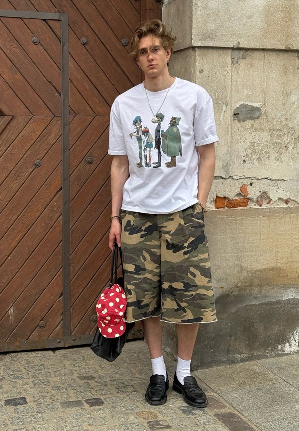 Jonge man met verwaaid haar staat tegen een muur, draagt een wit T-shirt met afbeelding, camouflage shorts, witte sokken en zwarte schoenen, en houdt een tas vast.