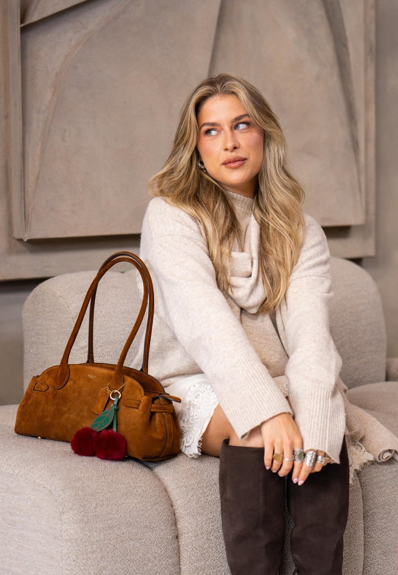 Braune Wildlederhandtasche mit doppelten Henkeln, verziert mit einem Anhänger mit grünem Blatt und zwei roten Bommel. Weicher beiger Pullover und kniehohe Stiefel.