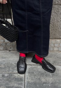 Persona che indossa scarpe in pelle intrecciata nere, calze rosse brillanti, una gonna di jeans scura, e tiene una borsa nera a rete su un pavimento di pietra.