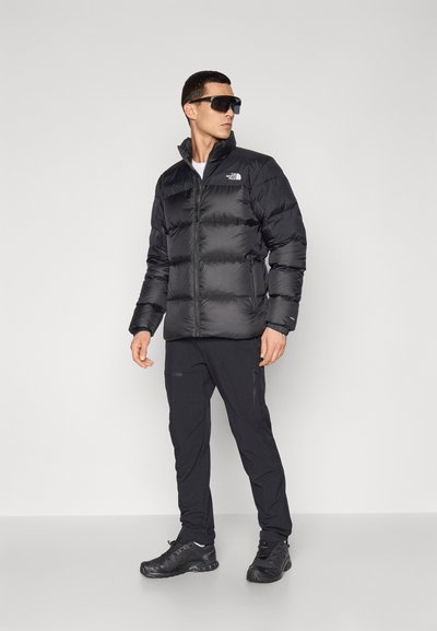 Chaqueta de plumas negra con cremallera en el frontal y cuello alto, con un logotipo en el lado izquierdo del pecho. Combinada con pantalones deportivos negros y zapatillas.