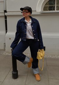Veste en denim avec deux poches avant, polo rayé, jeans évasés avec ourlets retroussés, sac à main jaune et bottes beiges. Casquette noire et lunettes.
