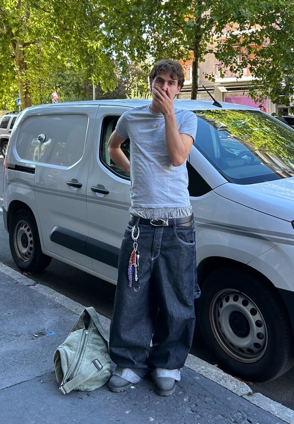 Weite dunkle Denim-Jeans, graues T-Shirt, schwarzer Gürtel mit Schlüsseln und eine grüne Stofftasche auf dem Boden. Im Hintergrund sind ein weißer Transporter und Bäume zu sehen.