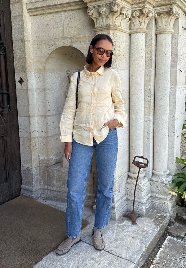 Femme aux cheveux courts et foncés portant des lunettes de soleil, une chemise beige à boutons, un jean bleu et des mocassins, debout devant un bâtiment à colonnes en pierre.