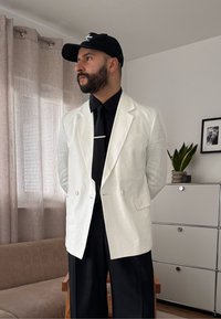 Homme barbu portant une casquette noire, un blazer blanc, une chemise noire et une cravate, se tenant dans une pièce avec un rideau beige, un canapé, une plante et une armoire.