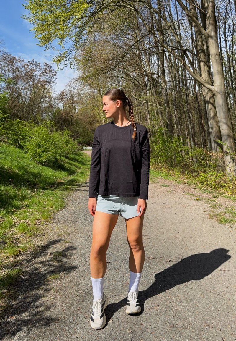 Femme en chemise noire à manches longues, short clair et baskets, debout sur un sentier forestier éclairé par le soleil, regardant vers sa gauche.