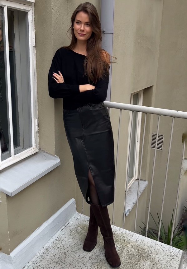 Pull en tricot noir, jupe en cuir noir taille haute avec une fente sur le côté, bottes en suède marron jusqu'au genou, se tenant sur un balcon.