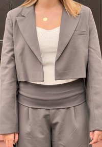 Kratka siva blazer jakna preko svetle teksturirane bluze s okruglim izrezom i sivim hlačama visokog struka. Ima čiste linije i bez vidljivih kopčanja.