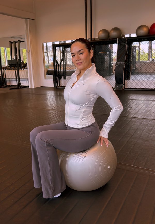 Une femme en tenue de sport est assise sur un grand ballon d’exercice dans une salle de sport, avec du matériel de fitness derrière elle.