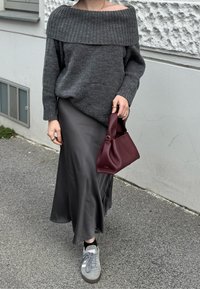 Person som bär en grå stickad off-shouldertröja, svart satin kjol, grå sneakers, håller en vinröd läderhandväska, går på en trottoar.