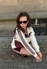 Balts un bordo sporta tērps ar lielām svītrām uz piedurknēm, tumši saulesbrilles, maza bordo krusta tipa soma, sēžot uz rakstaina akmens bruģa.