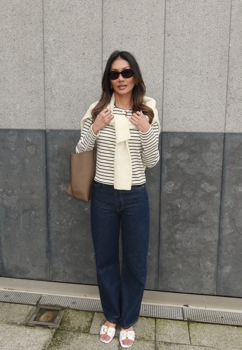 Camisa de manga longa às riscas, suéter creme nas ombros, jeans azul escuro e sandálias brancas com detalhes dourados. Bolsa tote castanha.