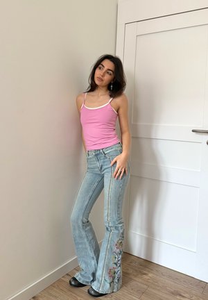 Joven mujer con camiseta sin mangas rosa y jeans azules con bordado floral apoyada contra una pared cerca de una puerta blanca cerrada, mirando de lado.